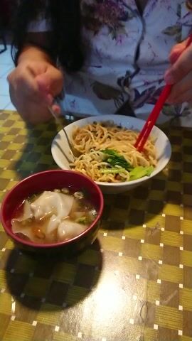 The video captures an overhead view of a meal being eaten at a table. The scene features a checkered tablecloth in shades of yellow and brown.

In the center, there are two bowls of food. To the left, a white oval bowl contains a serving of noodles mixed with green vegetables, possibly bok choy or a similar leafy green. A pair of red chopsticks is shown actively lifting some of these noodles from the bowl. To the right, a smaller, dark red bowl holds a clear soup with several white dumplings or wontons floating in it. A spoon is also placed within this red bowl.

A person's hands are visible in the frame. One hand, partially covered by a floral-patterned garment, holds the red chopsticks. Another hand, visible at the bottom left, holds the handle of the spoon in the red soup bowl. The focus is on the act of eating.

The setting appears to be indoors, possibly a home or a casual restaurant. The lighting casts a clear shadow from the red bowl onto the tablecloth, suggesting a light source from above. No specific location cues or text are visible in the frame.