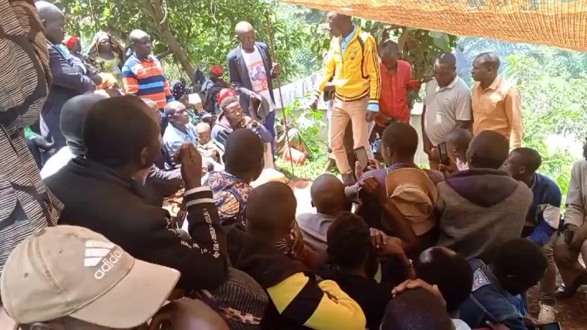 A group of about 30 people are gathered outdoors under a makeshift shelter. The shelter appears to be a sheet or tarp stretched between trees. Most people are seated on the ground, facing a few men who are standing. One man in a yellow jacket is gesturing and appears to be addressing the group. Another man standing near him is holding a walking stick. The people are a mix of ages and are dressed in casual clothing. Some people are holding phones and recording. The environment appears to be a rural or wooded area.