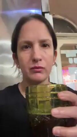 A woman is recording a video of herself indoors, drinking from a dark green glass. The glass has a raised design on the outside. She is wearing a black shirt, pearl earrings, and a gold necklace. She has dark hair pulled back and a neutral expression. Behind her is a stainless steel appliance, and in the background, a doorway and signs are visible.