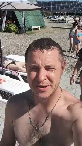 A man is taking a selfie outdoors on a sunny day. He is shirtless and wearing a gold chain. In the background, other people are visible. One woman is wearing a blue bikini, and another is wearing a black and white patterned cover-up and a black baseball cap. There are lounge chairs and solar panels in the background, suggesting a resort or beach area. The man is smiling at the camera. The video appears to be a casual personal recording.