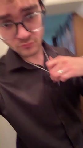 A man wearing glasses, a silver chain with a cross, a black button-up shirt, and black pants adjusts his black belt and looks at the camera. He is standing in a hallway with white walls, dark flooring and a wooden door frame on the right.