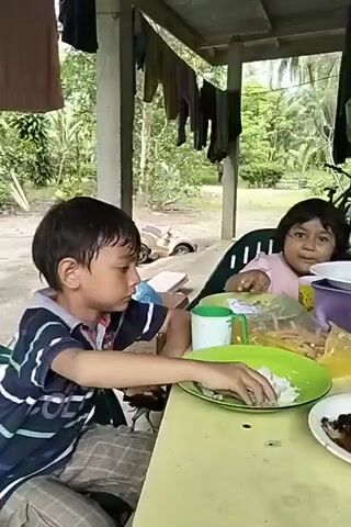 Two children are sitting at a table outdoors, eating a meal. The boy on the left is wearing a blue and white striped shirt and plaid shorts. He is looking to his right as he eats with his hands. The girl on the right is wearing a pink shirt and is also eating. In front of them on the yellow table are green plates with rice and what appears to be fish. There is also a mug, a plastic bag of long, thin snacks, and other dishes. A black chick walks near the boy's chair.  In the background, clothes hang from a line under a tin roof, with trees and a small vehicle visible in the distance.