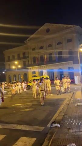 This video captures a nighttime street scene with a large, ornate building in the background, possibly a government or cultural institution, illuminated by exterior lights. A group of people dressed in white attire and red sashes are gathered in the street, some walking and others standing in conversation. A yellow ambulance is parked nearby. The street is paved, and a crosswalk is visible. In the middle ground and background, more people are seen, some sitting on a large monument or sculpture. The overall atmosphere appears to be a public gathering or celebration.