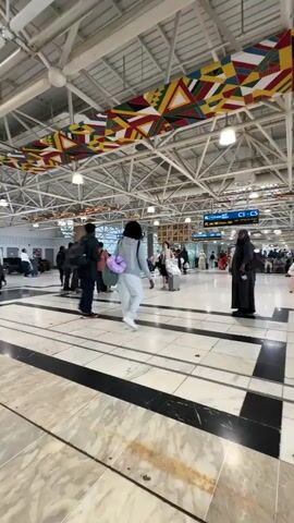 The video depicts a busy airport terminal with a high ceiling featuring exposed metal beams and decorative triangular banners in various colors. The floor is polished marble with black and white striped borders. Several people are walking through the terminal, some with luggage. In the foreground, a person in a dark jacket walks towards the camera, obscuring the view of the background momentarily. Other individuals are seen in the middle ground, walking in different directions and gathered in small groups. A sign with "C1-C5" is visible in the background, indicating gate numbers. The overall atmosphere suggests movement and travel.