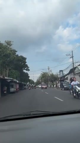 The video displays a street scene in Sumbersari, Indonesia. The perspective is from inside a moving vehicle, looking forward. The road is paved and appears to be a two-way street with multiple lanes. On the left side of the road, there are small shops or stalls with awnings, and several motorbikes parked. On the right side, there are buildings, some with signs, and more motorbikes, including one with two people riding it, a man in front and a woman behind him. There are also cars on the road, including a dark-colored SUV or similar vehicle.

The activity appears to be everyday street life, with vehicles moving along the road and people engaged in various activities related to the shops and transportation.

The time of day seems to be daytime, possibly late morning or afternoon, given the lighting. The weather appears to be overcast with clouds visible in the sky, but there is still enough light to see clearly. The presence of tropical trees on the left side of the road suggests a warm climate. There are no clearly discernible emotions visible in the individuals in the frame, as they are either distant or their faces are not clearly visible.

There is some text visible on signs on the right side of the road, but it is not legible due to the resolution and distance. One sign appears to be blue, and another has a white background with dark lettering. There is also a tall tower or antenna visible in the distance. The overall impression is a typical bustling street in a Southeast Asian town.