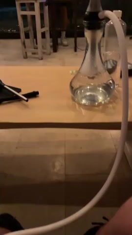 The video shows a hookah on a light brown wooden table, with the camera slowly panning up to reveal the room it's in. The hookah is gray and clear, with smoke inside the glass base. A white hose is attached to it. Behind the table, a person is DJing, using a laptop and mixer. They are standing in what appears to be a greenhouse or enclosed patio, with a glass roof and walls. String lights are visible along the ceiling, and the background is dark, suggesting it's night. A red Nike shoebox is on the stool where the DJ is working. A chessboard, bottle of water, and some miscellaneous items are on the table with the hookah.