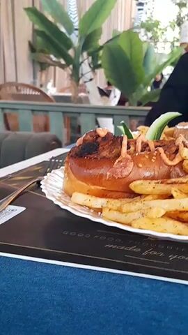 The video shows a close-up of a plate of food, likely a burger and fries, on a table in what appears to be a restaurant. The food is in focus in the foreground, with a long bun topped with meat and sauce, surrounded by golden-brown french fries. In the background, there are plants, chairs, and other tables, suggesting a dining setting. A person's arm and shoulder are visible in the upper right corner, and a fork is visible on the left side of the plate. The lighting is soft and natural, and the overall atmosphere seems relaxed. The video is shot from a slightly low angle, emphasizing the food.