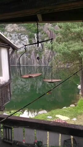 The video displays an outdoor scene from a slightly elevated vantage point, looking across a body of green water towards a steep, grey, rocky cliff face on the left and a lush green forested area on the right. In the foreground, a dark wooden railing extends horizontally across the bottom of the frame. Hanging from an unseen structure above, two large, circular, flat metal platforms are suspended by chains, forming a large, rustic balance scale. The left platform holds a small, light-brown, irregularly shaped object, causing it to sit slightly lower than the empty right platform. The water reflects the surrounding landscape, including the cliff and trees. No people are visible, and there is no discernible action or interaction. The camera remains static throughout the clip.