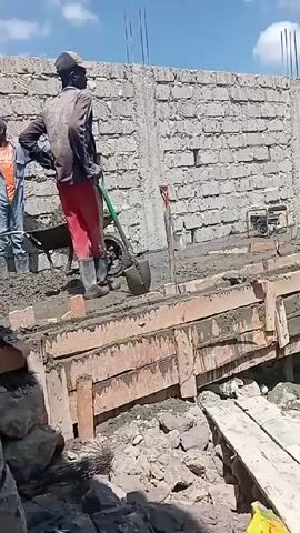 Construction workers are pouring concrete into wooden forms at a construction site. One worker is operating a machine, and several other workers are using shovels to distribute the concrete. A brick wall is in the background. The site is somewhat cluttered with building materials, rocks, and debris. The sky is partly cloudy.