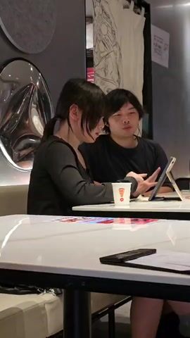 Two people, a woman with her hair in a ponytail and a man, are sitting at a table in what appears to be a cafe or restaurant. The woman is wearing a black long-sleeved shirt and the man is wearing a black t-shirt. They are both looking at a tablet device that is on the table in front of them. The woman is pointing at the tablet, and the man is also gesturing with his hands as if explaining something. On the table, there is a white cup with a red logo, a can of green soda, and some papers with colorful designs. In the background, there is a large circular mirror and some artwork on the wall. The overall atmosphere seems casual and engaged.