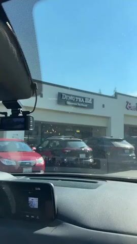 This is a view from the inside of a car, looking out through the windshield at a strip mall. The sky is blue and clear with a bright sun. In the foreground, the dashboard of the car is visible, with a digital display showing the time as 5:06. Through the windshield, several cars are parked in front of a row of storefronts. One storefront has a sign that reads "Ding Tea" and "Taiwanese Tea House". Other businesses visible are "Baja Fresh" and "Sport Clips". The scene appears to be a sunny day in a suburban area.