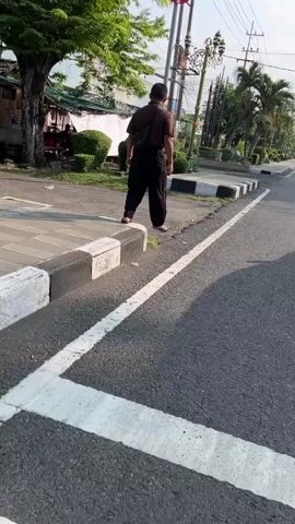 A person wearing a dark brown shirt, dark pants, and a black crossbody bag stands on a sidewalk next to a street. The person is facing away from the camera. In the background, there is a tree, some buildings, and utility poles. The ground is paved.