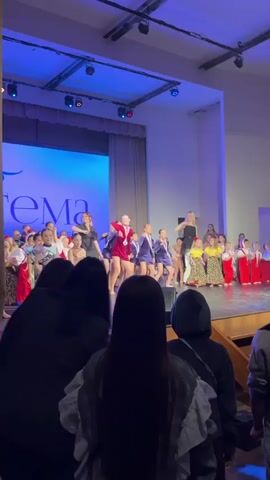 The video shows a group of children and adults standing on a stage, participating in what appears to be a dance performance or competition. The children are dressed in various costumes, including red, blue, and brown outfits, as well as patterned dresses. Some of them are wearing medals around their necks. There are two adults, who appear to be instructors, standing behind them. They are also moving to the music. The stage has a blue screen in the background with text on it. In the foreground, the backs of several people in the audience are visible. The lighting is bright and colorful, creating a cheerful atmosphere.