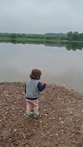 A young child stands on a rocky shore facing a calm lake. The child is wearing a dark blue vest over a blue long-sleeved shirt and light-colored pants with blue and green shoes. The child is holding up their right hand as if to throw something. In the background, across the lake, is a lush green treeline. To the right of the child, a gray inflatable boat with an outboard motor is partially visible, resting on the shore. The sky is overcast.