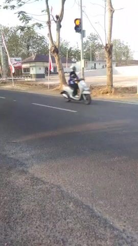 The video shows a street with a black car driving past on the right. There is a traffic light with a yellow light illuminated above the road. Two motorcycles are then seen traveling down the road. The environment appears to be a rural area with trees and buildings visible in the background. The weather is clear and the sun is shining, casting shadows on the road. There are flags on poles in the background, possibly indicating a national holiday or event.