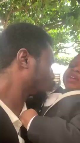 A man holding a small child dressed in a black suit and bow tie kisses the child's cheek. The child looks uncomfortable and makes a displeased sound. A partially visible tree is in the background. A man is partially visible in the background as well. He is wearing a white shirt and is holding something to his ear.