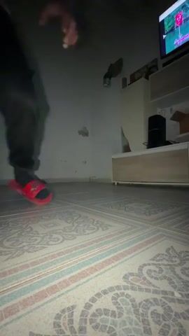 Here is a description of the video:
A person with long dark hair is breakdancing on a tiled floor in what appears to be a dimly lit living room. The person is wearing a black and pink long-sleeve shirt, black pants, and red slides. They perform a handstand-like move with their legs extended upwards, maintaining balance with their hands. In the background, a television is visible on a cabinet, displaying a sporting event. A light fixture hangs from the ceiling, and a decorative piece sits on top of the cabinet. The room is simply furnished, with the patterned tiled floor being a prominent feature.
