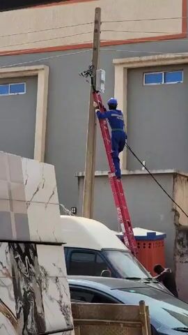 A medium shot captures a utility worker in a blue uniform and hardhat, positioned on a red ladder leaning against a utility pole. The worker appears to be engaged in some task related to equipment attached to the pole. A black wire is seen running from the pole. In the foreground, a white van with a dark blue cab is parked near the base of the ladder. To the left of the van, stacks of large, white marble-patterned tiles are leaning against the van. A man in a dark shirt is standing near the van, possibly assisting the worker. Behind the scene, a gray building is visible with evenly spaced windows and an orange accent line. A woman in a pink hijab is also present in the scene.