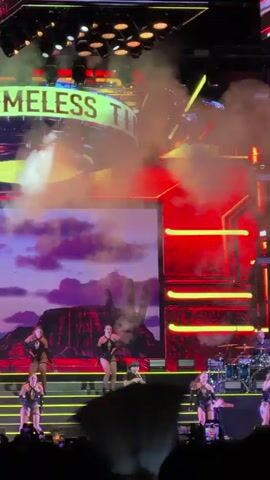 A video clip from a live performance captures a singer on stage, wearing a black hat and dark clothing, performing with a group of female dancers in revealing outfits. Behind them, a large screen displays a desert landscape with cacti and a purple sky. The stage is illuminated with bright lights, and smoke effects are visible. The words "TIMELESS" and "TIMBER" are displayed on a large screen at the top of the stage, suggesting the performance might be related to a song or tour. The audience is visible in the foreground, holding up phones to record the event. A drum set and other musical instruments are set up on stage, indicating a live band is present. The overall atmosphere is energetic and visually striking, characteristic of a large-scale concert.