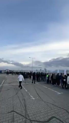 The video shows a crowd of people gathered in a large paved area, possibly a parking lot or an airport tarmac. The sky is partly cloudy. In the background, there are some ambulances, a red tent, and some buildings. People are walking around, some are standing in line. One person dressed in a unique black and yellow outfit walks by. The ground is paved with gray and white bricks and shows tire marks. There are light poles illuminating the area.