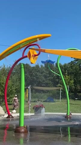 This video shows a children's splash pad on a sunny day. Brightly colored, curved structures with water sprayers are positioned over a wet, paved area. Two children and one adult are visible in the background. The adult is walking away from the camera on the left side, while one child is in the middle of the frame near a planter of flowers, and the other child is on the right. Trees and a clear blue sky form the backdrop.