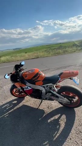 A motorcycle is parked on the side of a road in front of a field under a blue sky with white clouds. The motorcycle is orange and white with the Repsol logo visible. The field behind the motorcycle is green and rolling, with trees scattered throughout. The road appears to be asphalt, and the shoulder is gravel. The sun is casting shadows from the motorcycle onto the ground. The video appears to be shot from a stationary position, with the camera panning slightly to the left and then remaining mostly still.