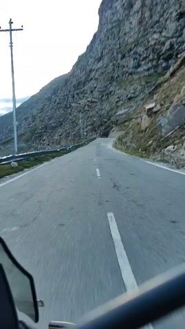A first-person perspective video shows a motorcycle traveling on a paved road next to a steep, rocky mountainside. The road has a white line dividing the lanes. There are utility poles along the side of the road. The sky is overcast. The motorcycle has a black windshield in the foreground.