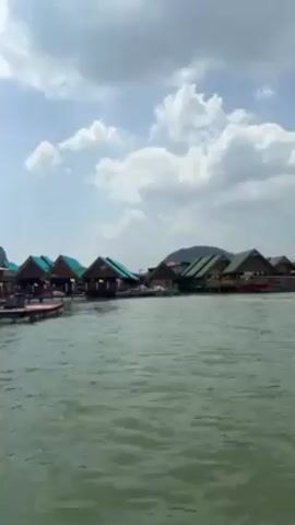The video shows a view from a boat traveling on calm waters, towards a village of stilt houses. The houses have brown walls and bright turquoise roofs.  A low mountain rises in the background. The sky is blue with scattered white clouds.