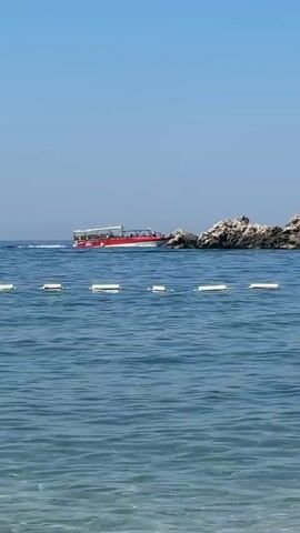 This video captures a scenic view of the Adriatic Sea, near Pržno, Montenegro. The primary subject is a red and white tour boat filled with people, cruising through the blue water under a clear, bright sky, indicating a sunny day, likely in the late morning or early afternoon. The boat is moving from left to right, passing by a rocky outcrop that juts out of the sea. The water is calm with gentle ripples, and a line of white buoys marks an area of the sea. The overall mood is peaceful and suggests a leisurely day of sightseeing or a boat tour. No visible text is present in the video.