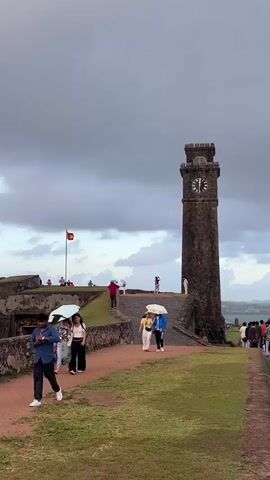 The video shows a view of a clock tower and people walking in what appears to be a historic fort. The sky is cloudy and the ground is a mix of grass and dirt pathways. In the foreground, a person in a blue shirt is walking away from the camera, while two people with umbrellas walk toward the camera. Further up a path, a couple holding an umbrella walks toward the clock tower. In the background, there are more people, a flag on a flagpole, and the stone ramparts of the fort. The clock tower is tall and made of dark stone with a clock face visible at the top. The overall atmosphere suggests a cloudy day with a few tourists exploring the site.