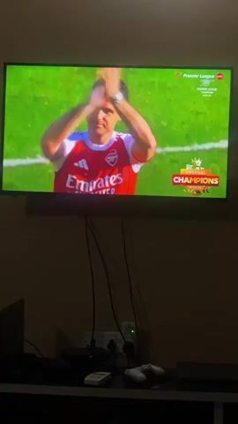 Here is a description of the video:

The video shows a person watching TV. The TV screen shows a man in a red and white Arsenal football jersey clapping. The screen also displays "Arsenal Champions." The room is dark, and a desk below the TV holds gaming consoles and controllers.  The camera is still.
