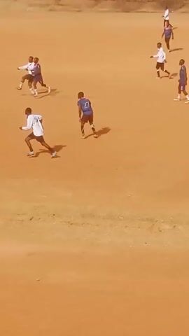 The video shows a soccer game in progress on a reddish-brown field, possibly dirt or clay. Several players are visible, some wearing white jerseys and others wearing blue and pink jerseys. The players are scattered across the field, running and kicking a soccer ball. One person in a bright yellow jersey and dark pants appears to be the referee, standing near a red bag. In the background, there is a tall concrete fence or wall.