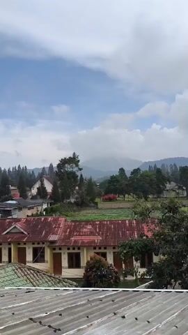 The video shows a medium shot of a village nestled in a valley, with rolling hills and mountains in the distance. The sky is partly cloudy, with a mix of blue sky and white clouds. In the foreground, there are several buildings with red tile roofs. Some of these buildings appear to be houses, while others might be small businesses or guesthouses. A dirt road or pathway is visible between the buildings, and some vegetation, including trees and small bushes, dots the landscape. In the middle ground, there's a cultivated field, possibly for agriculture, with rows of plants. The overall atmosphere is serene and rural, suggesting a peaceful, possibly mountainous, region. No people are visible in the frame.