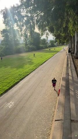 This media file captures a serene outdoor scene during what appears to be late afternoon or early evening, given the warm, angled sunlight and long shadows. The setting is a park or recreational area with a paved path suitable for cycling and running, bordered by a grassy field on one side and a tree-lined embankment on the other.

The primary subjects are individuals engaged in recreational activities. In the foreground, a person is cycling on the paved path, casting a long shadow. Further along the path, another person is running. In the background, several other individuals are scattered across the grassy field, some appearing to walk or stand, suggesting a leisurely atmosphere. Tall, slender trees with lush green foliage dominate the right side of the frame, with sunlight filtering through their leaves, creating a dappled effect. The grass in the field is vibrant green, indicating a pleasant climate.

The overall mood is one of peaceful activity and enjoyment of the outdoors. The lighting suggests a mild, likely warm, day. While the exact location within Sant Adrià de Besòs isn't explicitly identifiable through landmarks, the presence of a well-maintained path, open green spaces, and mature trees points to a public park or riverside trail designed for recreation. No visible text is present in the media.