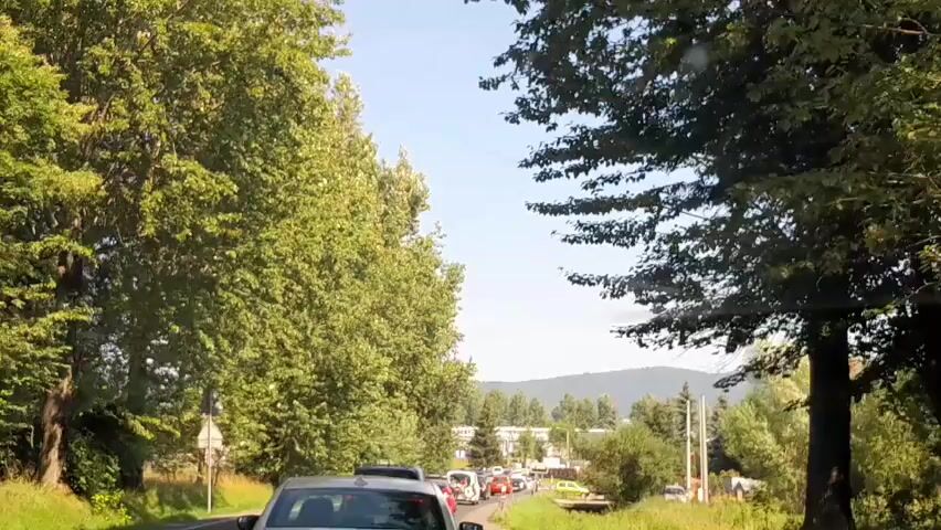 The video shows a view from inside a car driving on a road lined with trees. In the distance, there is a traffic jam with several cars. The sky is blue with some clouds, and there are mountains in the background. The sun appears to be shining. The video is taken in daylight.