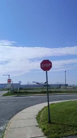 A passenger jetliner is captured in mid-flight, descending towards an airport runway. The aircraft, seen from a side profile, appears to be in the final stages of its approach. It is positioned against a backdrop of a clear blue sky dotted with scattered white clouds. In the foreground, a paved road curves gently, with a concrete sidewalk running alongside it. A prominent red octagonal stop sign stands on a metal pole on the right side of the road, indicating a traffic control point. Beyond the stop sign, a chain-link fence separates the road area from the airport grounds. A parking lot filled with cars is visible behind the fence, suggesting proximity to the airport terminal or a long-term parking facility. Streetlights are also present, contributing to the infrastructure of the airport environment. The sun is shining brightly, casting a lens flare into the upper left portion of the frame.