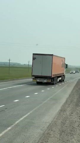 A semi-trailer truck is traveling on a highway. To the left of the highway, a tractor with a blade attachment is moving along the shoulder of the road. A white sedan overtakes the tractor, and then a black sedan passes the white sedan. The weather is overcast, and there are fields and utility poles in the background.