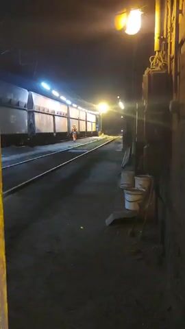 A nighttime scene at a train yard shows a worker in an orange vest near a train. The train is parked on a track, and the area is illuminated by overhead lights. There are several buckets and some tools leaning against a wall in the foreground. The ground is covered in dark, possibly dusty or sooty, material.
