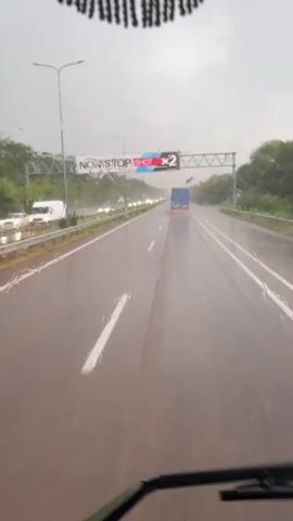A dashcam view shows a wet highway during rainfall. A blue truck is driving ahead, and other vehicles are visible in the distance. A sign above the highway reads "NON STOP SHOT X2". The windshield wipers are intermittently active. The scene appears to be in a rural or suburban area with trees lining the sides of the road. The sky is overcast and gray.