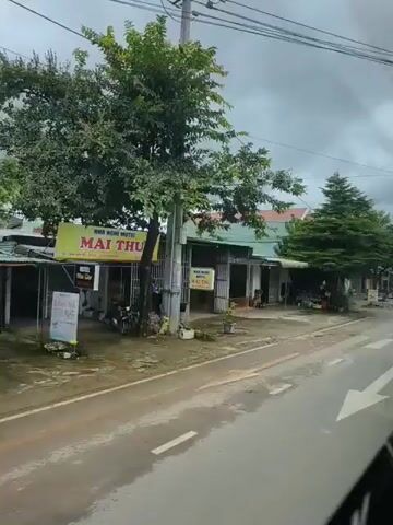 The video captures a street scene in what appears to be a rural or suburban area. A road with crosswalk markings runs through the center of the frame. On the left side of the road, there are several buildings with open fronts, resembling shops or small businesses. Some are simple structures with corrugated metal roofs, while others have more substantial facades and are painted in various colors. There are trees lining the street and in front of some of the buildings, some of which are quite large and leafy. The sky is overcast and the ground appears wet, suggesting recent rain.

Several objects are visible:
- A motorcycle is seen riding on the road in the foreground.
- In front of the shops, there are signs, some with text and images.
- There are various items of merchandise or stock stored outside some of the businesses, including what looks like barrels and what could be motorbikes parked.
- A few potted plants are placed outside one of the buildings.
- Power lines are strung overhead, typical of a populated area.
- In the distance, more buildings and trees can be seen, extending the streetscape.

The overall impression is that of a functional, everyday street in a community, with businesses operating and people using the road. There is no clear indication of any unusual activity.