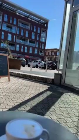 The video shows a cup of coffee on a table in the foreground with a view of a street and buildings in the background on a sunny day. The cup is white and sits on a saucer. Behind the table, the ground is paved with gray bricks. A chalkboard menu board is visible to the left. Across the street, there is a modern brick building with many windows. A few cars are parked along the street. A person on a bicycle rides past. A glass structure is to the right of the frame. The sky is clear and blue with a contrail visible.