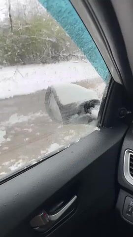 A car's interior is visible in this video clip. The view is from inside the vehicle looking out the driver’s side window. It is either raining or snowing, as water droplets and some ice are seen on the glass. The side mirror, covered in snow, is visible outside the window. The landscape outside is snowy, with trees.