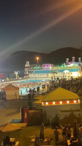 The video shows an aerial view of a crowded outdoor pool party at night. The pool area is decorated to resemble a cruise ship, with a long, multi-story structure mimicking a ship's deck and funnels. Numerous people are in the pool and surrounding areas. In the foreground, there are smaller structures with lights, picnic tables, and umbrellas, suggesting a recreational area. The sky is dark, with the scene illuminated by artificial lights from the pool area and some streetlights. Mountains are visible in the background.
