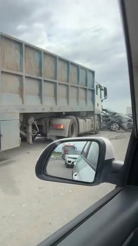 The video shows a car accident scene. A large, white truck is visible next to a heavily damaged gray Volkswagen Polo with a Russian license plate reading "T323OE 761". The front end of the car is crushed, the windshield is broken, and the hood is crumpled. One of the car doors is open. In the rearview mirror of the car recording the video, several other vehicles can be seen lined up behind, suggesting a traffic backup due to the accident. The road is asphalt and appears to have debris scattered across it. The sky is overcast.
