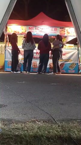 A group of five people, including a child being held by an adult, stand in front of a food stall at night. The stall is brightly lit and displays pictures of food items. The people are all facing away from the camera, towards the stall. One person is wearing striped pants and appears to be moving. The ground in front of the stall is paved, and there is some vegetation in the foreground. The overall scene suggests a casual outing or meal at a food vendor.