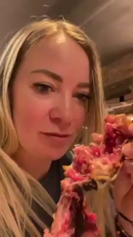 A fair-skinned woman with blonde hair and blue eyes holds up a large piece of meat and eats it in the video. Her hair is highlighted and styled in a center part, falling around her face. She is wearing a dark-colored shirt. Her fingernails are painted in light colors. The woman takes a bite of the meat, chews, and then looks at the meat in her hand, showing it to the camera. The meat is pink and brown, with some dark areas. The background includes a light-colored wall with brick detail and a glimpse of another person sitting at a table. The lighting appears to be from a mix of natural and artificial sources.