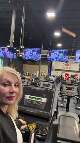The video opens with a blonde woman in a black and white athletic jacket taking a selfie in a gym. She is smiling and looking at the camera. Behind her, there are rows of treadmills, some of which are in use by other gym-goers. The gym appears to be well-lit with fluorescent lighting and has several televisions mounted on the walls displaying various programs. The woman then turns the camera slightly to show more of the gym environment, panning across the treadmills and the people on them. The video continues to show the woman interacting with the treadmill console, pressing buttons and adjusting settings. The background remains a blur of gym equipment and other patrons. The overall scene depicts a typical gym environment with individuals engaged in cardiovascular exercise.