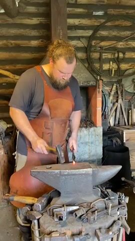 A blacksmith with a beard and hair in a bun, wearing a leather apron over a dark t-shirt, works at an anvil in a rustic workshop. He is using a hammer to shape a glowing piece of metal held in tongs. The workshop is made of logs, and various blacksmithing tools are visible in the background, including hammers on a wooden base beneath the anvil and a large metal wheel mounted on the wall. A brick forge is also present.