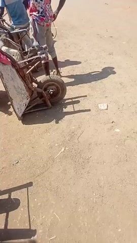 The video shows a person standing next to a small, beat-up cart with a single wheel in a dusty area that appears to be an outdoor market. The person is wearing sandals, light-colored pants, and a colorful shirt. The cart is old and appears to be made of metal and wood. The surrounding area is populated with people, cars, and other carts. A woman in a red dress is seated nearby, working with her hands. The ground is dirt and gravel, and there are tents or tarps set up to provide shade. The video appears to be taken during the day in sunny weather.