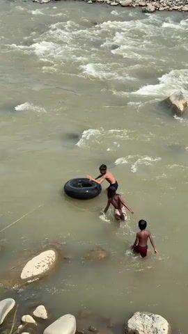 Three young boys are playing in a wide, shallow river. The water is murky and fast-flowing, with white rapids visible in the background and large rocks scattered throughout the riverbed. The boys are all wearing swim trunks. One boy is holding onto a large, black inner tube, seemingly trying to get it into position. Another boy is standing in the water next to him, reaching out towards the tube. The third boy is a little further away, standing in the water and looking towards the other two. The scene suggests a hot, sunny day, typical for a river in Seunong, Indonesia. The boys appear engaged in their play, their movements suggesting a playful or adventurous mood as they navigate the river and the inner tube.
